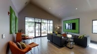 This single-floor space presents a well-designed open lounge area characterized by striking architectural details and thoughtfully curated furnishings. The most prominent architectural feature is the vaulted ceiling, which rises dramatically to create an expansive, airy atmosphere throughout the interior. Large floor-to-ceiling glass doors and windows along one wall flood the space with abundant natural light while offering direct views of a landscaped outdoor area and surrounding greenery.

The flooring transitions between light-toned tile near the entrance and glass doors, and warm wood-style flooring throughout the central and rear portions of the space. Seating is arranged in multiple distinct groupings, featuring dark charcoal sofas accented with green and patterned throw pillows, complemented by bold orange lounge chairs that add a vibrant pop of color throughout.

A standout decorative element is a vivid green textured wall panel framing a mounted flat-screen television, serving as a focal point along one wall. Recessed lighting in the ceiling supplements the natural light, ensuring the space remains well-lit. Toward the rear, a workstation or reception counter area is visible, suggesting a functional component to the otherwise lounge-oriented layout. Overall, the space conveys a modern, welcoming, and energetic ambiance.