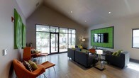 This single-floor space presents a well-designed open lounge area characterized by striking architectural details and thoughtfully curated furnishings. The most prominent architectural feature is the vaulted ceiling, which rises dramatically to create an expansive, airy atmosphere throughout the interior. Large floor-to-ceiling glass doors and windows along one wall flood the space with abundant natural light while offering direct views of a landscaped outdoor area and surrounding greenery.

The flooring transitions between light-toned tile near the entrance and glass doors, and warm wood-style flooring throughout the central and rear portions of the space. Seating is arranged in multiple distinct groupings, featuring dark charcoal sofas accented with green and patterned throw pillows, complemented by bold orange lounge chairs that add a vibrant pop of color throughout.

A standout decorative element is a vivid green textured wall panel framing a mounted flat-screen television, serving as a focal point along one wall. Recessed lighting in the ceiling supplements the natural light, ensuring the space remains well-lit. Toward the rear, a workstation or reception counter area is visible, suggesting a functional component to the otherwise lounge-oriented layout. Overall, the space conveys a modern, welcoming, and energetic ambiance.