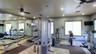 This fitness center offers a functional and well-appointed space designed to support a range of workout routines. The room features soft green-gray walls paired with recessed lighting and a ceiling fan, creating a comfortable and well-lit environment conducive to exercise. Large windows allow natural light to fill the space, contributing to an open and airy atmosphere.

The facility is equipped with a variety of cardio machines, including a treadmill, an elliptical trainer, and a stationary bike, catering to diverse fitness preferences. A multi-functional cable machine serves as the centerpiece for strength training, offering versatility for resistance-based workouts. Rubberized flooring mats are positioned beneath the equipment, providing stability and floor protection during use.

A wall-mounted television is visible near the treadmill, offering entertainment during cardio sessions. One full-length mirrored wall expands the perceived depth of the room while allowing users to monitor their form. The overall layout is organized and practical, making efficient use of the available space while maintaining an uncluttered, accessible environment for fitness activities.