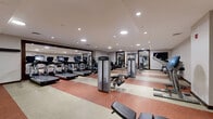 Located at 4200 North Carlin Springs Road in Arlington, VA, this fitness center offers a comprehensive workout facility designed to accommodate a variety of exercise routines. The space spans a generous single floor, providing ample room for both cardio and strength training activities without feeling cramped.

The layout is thoughtfully organized, with a dedicated cardio zone featuring multiple treadmills, elliptical trainers, and stationary bikes from Life Fitness — all positioned for comfortable use. Strategically placed large mirrors along the walls visually expand the space and allow users to monitor their form during workouts.

The strength training area is well-stocked with a selection of selectorized cable and resistance machines, a flat utility bench, and a dumbbell rack with a full range of free weights. A stability ball rounds out the functional fitness offerings. A wall-mounted flat-screen television adds an entertainment element to the space.

The flooring features a distinctive two-tone rubber surface in alternating red and beige stripes, providing both durability and traction. Recessed ceiling lighting delivers consistent, even illumination throughout the room. Near the entrance, a water fountain station and decorative wall art contribute a welcoming touch. The overall ambiance is clean, professional, and conducive to focused, effective exercise.