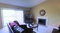 This single-floor space offers a warm and inviting atmosphere centered around a well-appointed living area. The living room serves as the heart of the home, featuring a tile-surround fireplace that adds both charm and functionality as a focal point. Large sliding glass doors dressed with vertical blinds flood the space with abundant natural light, creating an open and airy feel. The neutral, warm-toned walls provide a versatile backdrop that complements the room's dark leather seating and plush area rug. A classic round glass-top coffee table anchors the seating arrangement, adding a touch of elegance to the comfortable setting. Entertainment is well accommodated with dedicated media storage and display space positioned conveniently across from the seating area. The flooring transitions between soft carpeting in the main living zone and tile near the fireplace, offering both comfort and durability. Thoughtful details such as the decorative wall clock and subtle decor accents contribute to the space's lived-in, homey character. Overall, the layout is practical and cohesive, making it a functional and comfortable environment suited for both everyday relaxation and casual entertaining.