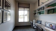 This thoughtfully designed single-floor space centers around a functional and stylish home office that exemplifies modern, purposeful design. The workspace is anchored by a built-in desk that runs along one wall, offering ample surface area for productivity, and is complemented by an ergonomic executive chair. Above the desk, open wall-mounted shelving provides organized storage and display space, currently styled with decorative accents, books, and greenery that add a touch of personality to the room. A large, black-framed window floods the office with natural light and frames a view of the surrounding outdoor landscape, creating an open and airy atmosphere. A tailored Roman shade in a warm taupe tone offers privacy when needed while maintaining the room's refined aesthetic. The walls are finished in a clean, neutral palette that enhances the sense of brightness, while the dark-toned flooring adds depth and contrast. The space also accommodates a printer and computer setup, making it fully equipped for a modern work-from-home environment. Overall, the space strikes a clean balance between practicality and contemporary style.
