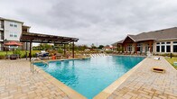 This expansive outdoor amenity area centers around a generously sized resort-style swimming pool with striking blue water and a paver stone deck that wraps around the perimeter. A standout feature is the gradual tanning ledge extending into the pool, fitted with in-water lounge chairs that allow residents to relax while partially submerged. The pool depth is clearly marked along the edges, and a water feature accent adds visual interest near one end.

The surrounding deck is finished with large-format stone pavers in warm tan tones, providing a clean and durable surface throughout. Numerous chaise lounge chairs are arranged along multiple sides of the pool, offering ample space for sunbathing. A covered pergola with a metal roof on one end of the deck provides shaded seating with outdoor dining furniture, while a second covered structure with a warm wood-paneled ceiling and substantial timber columns offers an additional lounge area with casual seating.

Adjacent to the pool sits a well-designed clubhouse building featuring stone and wood architectural elements, high peaked rooflines, and large windows that allow natural light inside. The grounds include manicured lawn areas, young ornamental trees, and landscaped beds that add greenery to the setting. The space is enclosed by a black metal fence, offering both security and open sightlines to the surrounding community.