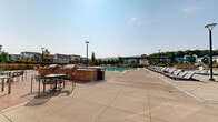This generously sized two-floor amenity complex offers an impressive range of recreational and social spaces designed for active community living. The exterior is defined by a contemporary façade combining natural stone cladding, crisp white panels, and floor-to-ceiling glass storefronts, with a second-floor balcony overlooking the outdoor grounds below.

At the heart of the outdoor area is a large, sparkling swimming pool surrounded by an expansive sun deck lined with modern lounge chairs, providing ample space for relaxation. A separate hot tub or spa area complements the pool, while a pergola-shaded zone adds a charming retreat from the sun. Hammock stands and fire pit seating areas are thoughtfully integrated into the landscaped grounds, which feature ornamental grasses, native plantings, and black iron fencing that frames open views of the surrounding greenery and neighborhood.

The patio dining terrace, accessible directly from the main building, accommodates multiple seating arrangements with green metal chairs and tables beneath a warm wood-accented overhang.

Inside, a soaring full-size basketball court commands attention, finished with hardwood flooring and decorated with bold "WE ARE" lettering and community branding. Industrial-height ceilings and large clerestory windows fill the space with natural light. A large roll-up door opens directly to the outdoor lawn, seamlessly connecting indoor and outdoor recreation.