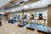 Located at 385 N Angier Ave NE in Atlanta, GA, this fitness center offers a comprehensive, single-floor layout designed to accommodate a wide range of workout routines. The main exercise area features an open, generously sized floor plan finished with neutral patterned carpet tiles, exposed ductwork along the ceiling, and large grid-paned windows that allow ample natural light to fill the space while offering views of the surrounding urban environment. Structural concrete columns add an industrial character to the room.

The cardio zone is outfitted with multiple treadmills and elliptical machines, each equipped with individual screens for entertainment. The strength training area includes a power rack, a punching bag, adjustable benches, and tiered dumbbell racks, as well as wall-mounted mirrors that enhance the sense of space. Ceiling fans and flush-mounted lighting ensure comfort throughout.

Two dedicated exercise studios are enclosed by sleek floor-to-ceiling glass partition walls with dark metal framing, providing visual openness while offering acoustic separation. One studio features hardwood-style flooring, wall-mounted televisions, stability balls, and a step platform for group fitness classes, while the other is set up as a cycling studio with multiple spin bikes and the same warm wood-toned flooring.