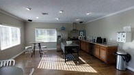 This single-floor residence offers an inviting, open-concept layout that seamlessly connects living and kitchen areas. The kitchen is a standout feature, showcasing warm honey-toned cabinetry, stainless steel appliances including a French door refrigerator with water dispenser, a gas range topped with a sleek wall-mounted stainless steel range hood, and a built-in dishwasher. A mobile kitchen island with a dark countertop provides flexible prep and serving space, while recessed lighting and a textured ceiling add character overhead. Crown molding runs along the ceiling perimeter, lending a polished, finished look to the space. Rich hardwood flooring extends throughout, catching natural light that pours in through a large window fitted with horizontal blinds, offering views of lush outdoor greenery. A casual dining area with a bistro-style table and stools sits comfortably near the window, creating a relaxed spot for everyday meals. Neutral gray walls create a versatile backdrop that complements both the warm wood tones and stainless steel finishes. The overall ambiance is bright, functional, and welcoming, making this a practical yet comfortable space suited for everyday living and entertaining alike.