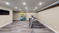 This fitness center offers a comprehensive workout environment designed to accommodate a variety of exercise routines. The space is finished with neutral beige walls, white baseboards, and striped, multi-tone carpet tiles that lend a clean and functional aesthetic throughout. Recessed lighting illuminates the entire area evenly, creating a bright and energizing atmosphere.

The cardio section features multiple Landice treadmills aligned along one wall, alongside a stair climber, providing ample options for aerobic training. Opposite this area, a dedicated strength training zone houses a multi-station cable machine and an adjustable weight bench, complemented by a well-organized dumbbell rack offering a wide range of free weights.

A functional training corner includes a floor-to-ceiling rack stocked with resistance bands, medicine balls, stability balls in assorted sizes, and rolled exercise mats, supporting stretching and dynamic workout routines. An exposed silver ductwork run along the upper wall adds an industrial accent to the otherwise polished interior. A wall-mounted flat-screen television provides entertainment during workouts. Two navy blue entry doors offer accessible points of entry, contributing to the room's overall organized and purposeful layout.