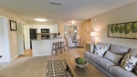 This single-floor residence offers an open-concept layout that seamlessly connects the kitchen, dining, and living areas, creating a cohesive and inviting atmosphere. The kitchen is equipped with black appliances, including a refrigerator and microwave, complemented by white cabinetry, a tile backsplash, and dark countertops. A breakfast bar with wooden bar stools provides a casual dining option and serves as a natural transition point between the kitchen and the rest of the living space. Adjacent to the kitchen, the dining area features an elegant table set surrounded by patterned upholstered chairs, accented by a decorative mirrored wall piece that adds depth and visual interest. The living room is anchored by a plush gray sofa adorned with colorful accent pillows, centered around a wooden coffee table atop a bold geometric-patterned area rug. Warm, neutral tones on the walls paired with soft carpeting throughout create a cozy and welcoming ambiance. Ceiling-mounted light fixtures provide consistent illumination across the open floor plan. The overall design balances comfort and functionality, making it an ideal space for both relaxing and entertaining.