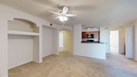 This single-floor, two-bedroom, one-bathroom residence offers a well-designed layout that maximizes both comfort and functionality. The living area is generously sized, featuring neutral-toned carpeting and a ceiling fan with integrated lighting, contributing to a bright and airy atmosphere. Built-in shelving niches are recessed into the walls, providing stylish display or storage opportunities without sacrificing floor space.

An elegant arched passageway connects the living area to the rest of the unit, adding a refined architectural detail that enhances the overall aesthetic. The kitchen is positioned adjacent to the living space in an open, pass-through configuration defined by a raised bar-height counter. It features warm wood-toned cabinetry, dark countertops, black appliances including a microwave and range, and track lighting overhead that ensures excellent task illumination.

The neutral color palette of white walls, light carpeting, and warm cabinetry creates a cohesive and inviting environment throughout. The single-floor layout ensures easy accessibility and seamless flow between spaces, making this a practical and comfortable residence.