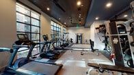 This modern fitness center is designed to support a comprehensive workout experience within a sleek, well-appointed single-floor layout. The space is defined by its dramatic floor-to-ceiling windows along one full wall, allowing abundant natural light to pour in while offering views of the surrounding outdoor environment — an energizing backdrop for any workout.

The cardio area is outfitted with a row of True-brand treadmills, elliptical trainers, and a stationary cycling bike, each equipped with individual display screens. A wall-mounted flat-screen television provides additional entertainment for those exercising in this zone. Overhead, a dark wood-paneled ceiling with recessed lighting and a ceiling fan creates a refined yet functional atmosphere, complemented by blue accent soffits that add a contemporary design touch.

The strength training section includes a cable crossover machine, a multi-functional cable station, and a dedicated TRX Training Zone complete with suspension trainers, a full dumbbell rack, medicine balls, and kettlebells — providing options for both free weight and functional training. The flooring features a patterned tile-carpet combination that delineates workout zones while maintaining visual cohesion throughout.

Overall, the fitness center balances modern design aesthetics with practical, high-quality equipment, creating an inviting and fully functional environment for a wide range of fitness activities.