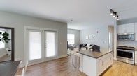 This single-floor residence offers a clean, contemporary design with an open-concept layout that seamlessly connects the kitchen and living areas. The kitchen features white cabinetry with modern hardware, a dark countertop island with bar seating, and a clean, minimalist aesthetic that anchors the space. Warm hardwood-style flooring extends throughout, adding a sense of continuity and warmth to the interior. The living area is generously proportioned, providing ample room for comfortable furnishings and everyday living. Large windows and a set of elegant French doors allow an abundance of natural light to pour into the space, creating a bright and airy atmosphere throughout the day. The neutral gray-toned walls complement the white trim and ceilings, lending the interior a fresh, modern feel. Recessed lighting and standard overhead fixtures provide well-distributed illumination across the open floor plan. The overall ambiance is light, welcoming, and functional, with thoughtful design choices that balance style and practicality. The open sightlines between the kitchen and living room make the space feel expansive and well-connected, ideal for both everyday living and entertaining.