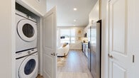Located on E 22nd Ave in Aurora, CO, this efficiently designed one-bedroom, one-bathroom unit offers a thoughtfully organized layout across a single floor. The open-concept kitchen and living area forms the heart of the space, where light wood-tone flooring, recessed lighting, and neutral walls create a clean, contemporary atmosphere. The kitchen is outfitted with full stainless steel appliances—including a French door refrigerator, electric range, built-in microwave, and dishwasher—complemented by white shaker-style cabinetry extending to the ceiling, granite countertops, a stainless farmhouse sink, and a white subway tile backsplash. The living area flows naturally toward a glass door that opens onto a private balcony, framed by large windows that bring in abundant natural light and offer open city views. A stacked washer and dryer unit is conveniently integrated near the entry corridor. The bedroom features carpeted flooring, a ceiling fan, and a generously sized window with additional city views, providing a comfortable and well-lit retreat. The bathroom includes a granite-topped white vanity, an illuminated frameless mirror, and wood-tone flooring, maintaining the unit's consistent modern aesthetic throughout.