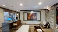 This single-floor space presents a well-designed contemporary interior that blends functionality with modern aesthetics. The main area serves as a lounge or lobby, anchored by a large framed wall-mounted display set within a bold, decorative accent wall featuring a contrasting dark trim border — a clear focal point of the room. Recessed ceiling lighting combined with pendant lights provides a layered, warm illumination throughout the space. The flooring features a multi-toned patterned carpet that adds texture and visual interest underfoot. Seating options include a cushioned bench, a pair of modern accent chairs, and bar-height stools positioned along a built-in counter or workstation area, suggesting the space may serve a dual purpose for both relaxation and productivity. A sleek dark-finished counter with a computer monitor reinforces a functional, service-oriented zone, potentially indicating a front desk or business station. An exposed brick accent wall adds an industrial yet warm character to one side of the room, complementing the neutral tones of the surrounding walls and ceiling. Overall, the space conveys a polished, inviting atmosphere suited for shared or communal use.