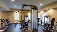 This dedicated fitness center is designed to support a comprehensive workout experience within a single-floor layout. The space is outfitted with professional-grade Life Fitness strength training equipment, including a cable pulley machine, a multi-function weight station, and a weight bench, providing users with a wide range of resistance training options. Cardio equipment, including a treadmill, is positioned toward the perimeter of the room, making efficient use of the available floor space. The flooring is a durable dark rubber material, purpose-built for high-traffic exercise environments and designed to absorb impact. Warm, neutral-toned walls contribute to an energizing yet comfortable atmosphere, complemented by crown molding details that lend a refined aesthetic to the space. Recessed ceiling lights provide consistent, even illumination throughout, while multiple white-framed windows along the walls allow natural light to filter in, offering views of surrounding greenery. A large wall-mounted mirror enhances the sense of openness and allows users to monitor their form during workouts. The overall design balances functionality with a clean, well-maintained ambiance suited for regular fitness use.