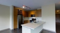 This single-floor space showcases a thoughtfully designed open-concept kitchen that serves as the centerpiece of the layout. The kitchen is outfitted with stainless steel appliances, including a full-size refrigerator, electric range, and over-the-range microwave, all seamlessly integrated into the design. Warm, medium-toned flat-panel cabinetry lines the walls, offering generous storage, and is complemented by a standout built-in diamond-pattern wine rack that adds a distinctive decorative touch. The countertops are finished in crisp white, extending to a sleek kitchen island that provides additional workspace and casual seating potential. The island also features an undermount sink with a modern arc-neck faucet. Dark hardwood flooring runs throughout, creating a rich contrast against the lighter walls and countertops. Track lighting mounted on the ceiling provides focused illumination across the kitchen workspace. The overall aesthetic is contemporary and clean, combining functional design elements with warm material choices to create a space that is both practical and visually inviting. The open layout promotes a sense of spaciousness, making this kitchen well-suited for both everyday living and entertaining.