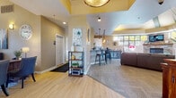 This single-floor community clubhouse is thoughtfully designed to offer a warm and functional gathering space. The open layout flows seamlessly from the entry area through the main lounge and into supporting spaces, creating an inviting and well-connected environment throughout.

The entry area features light wood-look flooring, a decorative pendant light with a distinctive tray ceiling accent in yellow, and an open shelving unit that adds a welcoming touch. Adjacent to the entry is a private office area highlighted by large windows that overlook an outdoor courtyard, filling the space with natural light. A substantial wood desk paired with upholstered chairs makes this area both professional and comfortable.

The kitchenette is outfitted with white upper cabinetry, a subway tile backsplash, sleek quartz countertops, and a raised bar with industrial-style metal stools. Warm Edison-style pendant lights hang above, lending the area a relaxed yet refined atmosphere.

The main lounge area is a standout feature, showcasing a floor-to-ceiling stacked stone fireplace flanked by large multi-pane windows that bring in abundant natural light and views of mature trees outside. Tray ceilings with bold yellow accents add architectural interest overhead, while plush carpeting and comfortable seating arrangements create a cozy, community-centered retreat.