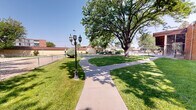 Located at 1177 Cardenas Dr SE in Albuquerque, New Mexico, this single-level community outdoor space offers a thoughtfully landscaped environment designed for leisure and recreation. The grounds are characterized by expansive, well-kept green lawns shaded by large, mature trees that provide natural relief from the Southwestern sun. Curving concrete walkways wind throughout the property, connecting various activity areas while classic multi-lamp street-style light posts add a charming, decorative touch along the paths.

A generously sized swimming pool serves as a central outdoor amenity, featuring clear turquoise water, a surrounding concrete deck, and seating areas with chairs and loungers for relaxation. The pool area is enclosed by an iron fence, enhancing safety and defining the space. Decorative rock landscaping and ornamental plantings add visual interest throughout the grounds.

The multi-story residential building adjacent to the grounds features balconies and brick and stucco exterior detailing, while a separate brick structure adds architectural variety to the campus. Picnic tables are positioned within the green areas, offering communal gathering spots. Overall, the space presents a welcoming, park-like atmosphere enhanced by abundant natural greenery and open New Mexico skies.