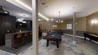 This single-floor amenity space is thoughtfully designed to deliver a upscale recreational and social experience. The layout is anchored by a full-size billiards table positioned beneath an elegant multi-arm chandelier, creating a focal point that draws attention immediately upon entry. Plush lounge seating is arranged nearby, offering a comfortable spot for guests to gather and unwind. The flooring features a patterned area rug that adds texture and visual interest to the space, complementing the neutral, warm-toned walls accented with framed artwork. Tray ceilings with integrated LED cove lighting add depth and a refined ambiance to the room, while recessed lighting throughout ensures the space is well-illuminated. Adjacent to the lounge area is a sleek bar section complete with counter-height seating and pendant lighting, finished in dark tones that contrast stylishly with the brighter lounge area. The overall design blends modern and contemporary elements, creating an inviting atmosphere well-suited for both casual socializing and organized events. The cohesive aesthetic, layered lighting, and purposeful furnishings make this a well-appointed communal amenity space.
