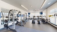Located at 15000 Penmon Dr in Fuquay-Varina, NC, this single-floor fitness center offers a generously sized, thoughtfully designed space for a comprehensive workout experience. The interior features a clean, modern aesthetic with white walls accented by a distinctive blue horizontal stripe, complemented by durable rubberized flooring in a speckled gray finish that transitions to a smooth surface in the functional training zone.

The facility is organized into distinct workout areas. The cardio section is lined with treadmills and Cybex elliptical machines positioned along large windows, flooding the space with natural light and providing open outdoor views of the surrounding neighborhood. Ceiling fans overhead ensure air circulation throughout.

The strength training area is anchored by a multi-functional cable and Smith machine system, a power rack with a bench press setup, and a full dumbbell rack offering a range of weights. Large wall-mounted mirrors enhance the sense of space and allow users to monitor their form. A dedicated free weights and functional training corner features stability balls, a plyo box, and additional floor space for stretching or bodyweight exercises.

Multiple wall-mounted flat-screen televisions are positioned throughout the facility for entertainment during workouts. Linear LED lighting provides bright, even illumination across the entire space, creating an energizing and welcoming environment well-suited for all fitness levels.