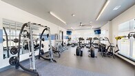Located at 15000 Penmon Dr in Fuquay-Varina, NC, this single-floor fitness center offers a generously sized, thoughtfully designed space for a comprehensive workout experience. The interior features a clean, modern aesthetic with white walls accented by a distinctive blue horizontal stripe, complemented by durable rubberized flooring in a speckled gray finish that transitions to a smooth surface in the functional training zone.

The facility is organized into distinct workout areas. The cardio section is lined with treadmills and Cybex elliptical machines positioned along large windows, flooding the space with natural light and providing open outdoor views of the surrounding neighborhood. Ceiling fans overhead ensure air circulation throughout.

The strength training area is anchored by a multi-functional cable and Smith machine system, a power rack with a bench press setup, and a full dumbbell rack offering a range of weights. Large wall-mounted mirrors enhance the sense of space and allow users to monitor their form. A dedicated free weights and functional training corner features stability balls, a plyo box, and additional floor space for stretching or bodyweight exercises.

Multiple wall-mounted flat-screen televisions are positioned throughout the facility for entertainment during workouts. Linear LED lighting provides bright, even illumination across the entire space, creating an energizing and welcoming environment well-suited for all fitness levels.