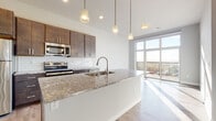 Located at 5909 Sharpsburg Dr in Madison, Wisconsin, this single-floor unit offers a clean, contemporary design with an open-concept layout that maximizes both functionality and natural light. The kitchen serves as the centerpiece of the space, anchored by a generous island topped with granite countertops and an undermount sink illuminated by a trio of pendant lights. Rich dark-toned cabinetry pairs with a classic white subway tile backsplash, while a full suite of stainless steel appliances — including a range, microwave, and refrigerator — completes the well-appointed cooking space. Light wood-tone flooring extends seamlessly throughout the unit, connecting the kitchen to the open living area. Floor-to-ceiling windows and a sliding glass door bathe the living space in natural light and provide direct access to a private balcony with outdoor views. The bedroom is bright and airy, benefiting from the same generous glazing and balcony access. The bathroom features granite countertops consistent with the kitchen, dark cabinetry with brushed nickel hardware, a large mirror, and a tub-shower combination, all finished with the same light flooring. The overall ambiance is modern, bright, and well-coordinated throughout.