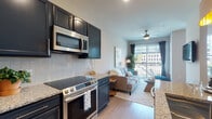 Located at 265 E State St in the heart of Columbus, Ohio, this well-appointed unit offers a thoughtfully designed single-floor layout that blends modern style with everyday functionality. The open-concept kitchen and living area form the heart of the space, where dark navy cabinetry, granite countertops, and a full suite of stainless steel appliances — including a refrigerator, electric range, and over-the-range microwave — create a sleek, contemporary kitchen. A geometric herringbone tile backsplash adds a refined design detail. The kitchen flows seamlessly into the living area, which features light wood-style flooring, a ceiling fan, and large windows with access to a private balcony overlooking the surrounding cityscape, filling the space with natural light. The bedroom is a calm and comfortable retreat, with soft neutral tones, carpeted flooring, and ample room for a full bedroom set. Adjacent, the bathroom is finished with matching granite countertops, dark cabinetry, a single vanity with a large mirror, and a glass-enclosed walk-in shower with light tile flooring throughout. A dedicated in-unit laundry area houses a stacked washer and dryer with additional wire shelving for storage. The overall aesthetic is modern and cohesive, with consistent finishes across all spaces.