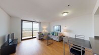 This high-rise unit offers a thoughtfully designed single-floor layout that balances comfort and functionality. The open-concept living and dining area is anchored by warm hardwood flooring, large sliding glass doors, and access to a private balcony that frames sweeping city and treetop views. The living space accommodates a sofa, accent chair, and dining table, creating a versatile area suited for both relaxing and entertaining.

The kitchen is outfitted with stainless steel appliances, including a full-size refrigerator, electric range, and built-in microwave, all complemented by clean white cabinetry and dark countertops. Natural light filters in through the kitchen's hardwood-floored entry.

The bedroom features plush carpeting, a full-size bed with coordinated nightstands, and a corner window arrangement that offers additional city views and abundant natural light. A dedicated workspace within the bedroom includes a desk, ergonomic chair, and dresser, making it well-suited for remote work.

The bathroom is finished with white tile and includes a full soaking tub with shower combination. Throughout the unit, a neutral color palette of whites and grays creates a calm, cohesive atmosphere. The overall design is modern and practical, well-suited for both short and extended stays.