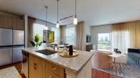 This well-appointed single-floor residence showcases a thoughtfully designed open-concept layout that seamlessly connects the kitchen, dining, and living areas. The kitchen serves as a focal point, featuring a large granite-topped island with an undermount sink, warm wood cabinetry with gold hardware, and pendant lighting overhead on a track rail system. Stainless steel appliances, including a full-size refrigerator and dishwasher, complement the subway tile backsplash, while hardwood flooring runs throughout the main living areas.

The living room is bright and welcoming, bathed in natural light from large windows dressed with sheer patterned curtains and horizontal blinds. A sliding glass door provides direct access to a private balcony with outdoor views, reinforcing the connection between interior and exterior spaces.

The bedroom offers a comfortable retreat, featuring multiple windows, a ceiling fan, and ample natural light. A generously sized walk-in closet is outfitted with built-in shelving, corner shelving units, and hanging rods, providing excellent organization potential.

The entryway, visible from the kitchen, features warm hardwood flooring and clean white walls, establishing a welcoming arrival experience. Throughout, the space maintains a cohesive, contemporary aesthetic with neutral tones and modern finishes.