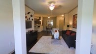 This single-floor residence features an open-concept design that integrates the living, dining, and kitchen areas into one cohesive, functional space. Dark hardwood flooring runs throughout, providing a rich, grounding contrast to the lighter walls and furnishings.

The living area is anchored by a dark sofa accented with bold red throw pillows, complemented by a plush white area rug and a sleek glass coffee table. A wall-mounted flat-screen television sits atop a low-profile, dark media console, while decorative wall art and sculptural accents add personality to the space. A ceiling fan with a light fixture provides both ambient lighting and airflow.

Adjacent to the living area, the dining space accommodates a compact dining table and chairs, positioned near a decorative mirror that reflects light and adds visual depth to the room.

The kitchen, visible from the main living space, features dark cabinetry, a breakfast bar with bar stools, and pendant lighting, making it ideal for casual dining and entertaining. A hallway visible toward the rear of the space leads to additional rooms, suggesting a well-organized and practical floor plan throughout.
