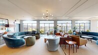 Located along W Winnemac Ave in Chicago's Ravenswood neighborhood, this single-floor lounge space combines contemporary architecture with a warm, mid-century modern aesthetic. Large floor-to-ceiling windows line the perimeter, flooding the interior with natural light and offering expansive views of the surrounding cityscape and elevated rail corridor.

The space is thoughtfully divided into distinct seating zones, each with its own character. A central lounge area anchors the room with a bold blue curved sectional sofa, complemented by swivel chairs in muted greens, teal, and cognac leather. A sculptural sputnik chandelier in brass serves as a focal point overhead, adding a retro-modern touch. Nearby, clusters of chartreuse accent chairs surround white Tulip-style tables, creating intimate conversation areas bathed in sunlight.

Polished concrete floors transition seamlessly to plush area rugs, delineating the various zones while adding texture and warmth. Lush potted plants are placed throughout, enhancing the welcoming ambiance. A built-in service counter with open shelving offers a functional utility area near the entrance corridor, which features additional seating and framed wall art. The overall atmosphere is bright, airy, and inviting.