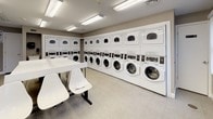 The Laundry and Fitness Center at 143 Shore in Peabody, Massachusetts is a well-maintained, single-floor communal facility designed for convenience and functionality. The laundry room is the centerpiece of the space, featuring an extensive row of commercial-grade front-loading washing machines paired with stacked dryers lining the walls on multiple sides. The machines are uniformly white and arranged in an organized, efficient layout that maximizes the use of the available floor space. Bright fluorescent lighting illuminates the entire room evenly, creating a clean and welcoming environment. The floor is finished with light-toned tile, contributing to the overall bright and tidy aesthetic. A large white folding table with modern hairpin-style legs occupies the center of the room, accompanied by white molded chairs — providing a practical surface for sorting and folding laundry. A digital kiosk near the entrance offers convenient access to laundry management and payment services. Informational signage and a fire safety panel are clearly visible along the walls. A glass-paned entry door allows natural light to filter in and offers a view to the exterior parking area. The space also features a utility sink and soap dispenser near a secondary door, adding to the room's practicality. Overall, this facility is clean, functional, and thoughtfully arranged.