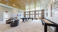 Located at 2300 SW 38th Ave in Ocala, Florida, this single-floor community living and game room offers a well-appointed gathering space designed for both relaxation and recreation. The room immediately impresses with its soaring ceilings finished in warm, natural wood planking, complemented by recessed lighting and an elegant pendant chandelier that anchors the central game area.

Large, floor-to-ceiling windows line one wall, dressed with neutral linen drapes that frame generous views of the lush outdoor greenery, flooding the interior with abundant natural light. The open layout is anchored by plush, patterned carpeting throughout, lending a comfortable and cohesive feel to the space.

The game area takes center stage with a full-size pool table and a foosball table, making it a focal point for social activity. A wall-mounted flat-screen television is positioned in the lounge zone, which features contemporary curved accent chairs arranged around a coffee table — ideal for casual gatherings.

An adjacent wall showcases a distinctive grid-panel wainscoting detail in soft gray and white, adorned with framed artwork that adds a refined, curated touch. A high-top table and bar seating provide an additional nook for conversation or casual dining. The overall aesthetic is modern and inviting, blending sophisticated design elements with functional, community-centered amenities.