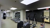 This fitness center is a dedicated, single-floor exercise facility designed to support a comprehensive cardio workout routine. The space is outfitted with multiple treadmills aligned along one wall, accompanied by elliptical cross-trainers and a stationary bike, providing a well-rounded selection of cardiovascular equipment. A large mirror spans one wall, visually expanding the room and allowing users to monitor their form during workouts. The ceiling features bright, evenly spaced recessed panel lights that flood the space with clean, consistent illumination, creating an energetic and functional atmosphere. Structural columns punctuate the floor plan, adding to the defined layout of the room. The walls are finished in a deep, dark tone on one side, contrasting with lighter finishes elsewhere, lending the space a modern and focused aesthetic. The flooring is a neutral, durable surface well-suited for fitness use. Overall, the fitness center offers a practical, no-frills environment that prioritizes functionality and ease of use, making it an ideal space for daily exercise routines.