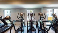 This single-level fitness center offers a thoroughly equipped workout environment designed to accommodate a wide range of exercise needs. The space features light hardwood-style flooring throughout, contributing to a clean and contemporary aesthetic that complements the neutral-toned walls and recessed lighting overhead. A distinctive wood-paneled ceiling with ceiling fans adds architectural character while maintaining airflow throughout the room.

The cardio zone is prominently lined with Matrix-brand treadmills, elliptical trainers, and stationary bikes, each equipped with individual digital display consoles. Large windows along the perimeter allow natural light to filter in, with views of an outdoor pool area and surrounding greenery providing a pleasant backdrop during workouts.

The strength-training section includes a cable and pulley functional trainer, weight benches, a dumbbell rack, and various resistance machines. A dedicated functional fitness area houses medicine ball racks, kettlebell sets, TRX suspension trainers, and a heavy punching bag, offering versatility for dynamic training routines. Full-length mirrors along one wall enhance the sense of space and allow users to monitor their form. The overall ambiance is modern, well-lit, and organized, creating an inviting environment for focused fitness activity.