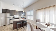 This single-floor residence is designed with a contemporary aesthetic, combining functionality with modern finishes throughout. The open-concept kitchen and dining area serves as the heart of the space, featuring dark espresso cabinetry, granite-style countertops, and a full suite of stainless steel appliances including a refrigerator, microwave, and range. A peninsula island with bar seating provides additional prep and dining space, while pendant lighting and a track lighting system illuminate the area effectively. The soaring ceilings amplify the sense of openness, extending through the main living areas. Light wood-look plank flooring runs through the kitchen, dining, and entry areas, while plush carpeting is found in the bedrooms and secondary living space. The dining area benefits from large windows dressed with sheer linen curtains, allowing generous natural light. A secondary carpeted room offers flexible use as a living area or additional bedroom. The bathrooms are consistently finished with dark cabinetry, granite-topped vanities, wood-look flooring, and framed mirrors. The bedrooms are comfortably scaled with bright windows and neutral tones, contributing to the unit's calm, cohesive atmosphere throughout.