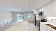 Located at 265 Reynolds Street in Oakville, Ontario, this single-floor unit presents a clean, contemporary aesthetic throughout. The open-concept layout connects the kitchen and living area seamlessly, creating a bright and functional living space enhanced by recessed pot lighting and light wood-tone flooring that runs consistently throughout.

The kitchen is a standout feature, equipped with sleek, high-gloss white cabinetry that extends to the ceiling, providing generous storage. A striking marble-look quartz island serves as a central gathering point and workspace. Stainless steel appliances — including a refrigerator, electric range, built-in microwave, and dishwasher — complete the modern kitchen ensemble. A window above the sink allows natural light to filter in.

The living area benefits from a sliding glass door that opens onto a private balcony, offering views of mature trees and green surroundings. An additional window further enhances natural light in the space.

The bedrooms are finished in soft neutral tones, with light wood flooring, roller blinds, and baseboard heating. At least one bedroom includes a sliding-door closet with built-in shelving. The unit's overall palette — crisp whites, warm beiges, and soft greys — contributes to a fresh, move-in ready ambiance throughout.