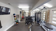 This well-appointed fitness center at 5201 Hunters Ridge Rd in Fort Worth, Texas, offers a comprehensive workout environment designed to accommodate a wide range of exercise routines. The space is thoughtfully laid out across a single floor, combining strength and cardio zones for a complete training experience.

The cardio area is bathed in natural light, courtesy of large windows that line the perimeter and provide open views of the surrounding outdoor grounds, including a pool area and lush greenery. Treadmills, elliptical trainers, and stationary bikes are positioned to take full advantage of these views, creating an energizing atmosphere for endurance workouts.

The strength training section features a full dumbbell rack with a wide range of weights, a multi-function cable machine, and a medicine ball rack with an assortment of weighted balls for functional training. Floor-to-ceiling mirrors are mounted along the walls, visually expanding the space and allowing for proper form checks during workouts.

Exposed brick columns add a warm, architectural character to the otherwise clean and modern interior, complemented by durable rubber flooring throughout. Overhead fluorescent lighting ensures the space remains bright at all hours. A water fountain is conveniently accessible within the facility, and vending machines are situated near the entrance for added convenience. Posted exercise room rules ensure a safe and orderly environment for all users.