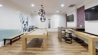 Located at 1001 Starbuck St in Fullerton, California, this single-floor amenity space is thoughtfully designed to serve as a social hub with a clean, contemporary aesthetic throughout. Warm wood-look flooring and bright white walls create a cohesive, welcoming atmosphere across all areas.

The dining lounge is a standout feature, offering multiple white square tables paired with chartreuse green modern chairs. Natural light filters in through windows, and a vibrant abstract painting anchors one wall, adding an artistic focal point. A wall-mounted flat-screen television provides entertainment, making the space equally suited for casual gatherings or communal relaxation.

Adjacent to the dining area is a well-appointed kitchenette, featuring dual countertops with ample cabinetry in a warm wood finish topped with white quartz surfaces. A deep stainless steel sink, pendant lighting, and decorative accents — including a bold gold "eat" wall letter — give this prep area both function and personality. Bar-height seating with patterned upholstered stools adds a casual dining option.

The game room is a lively centerpiece, highlighted by a full-size pool table beneath a striking double-orb pendant light fixture. A large-scale French Bulldog mural adds a playful, artistic energy, while a high-top bar counter with stools and a mounted television complete the entertainment-ready atmosphere. The layout flows intuitively between all zones, making this a versatile and engaging communal retreat.