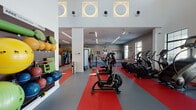 This spacious fitness center is designed with both functionality and aesthetics in mind, offering a comprehensive workout environment across a single open floor plan. The interior is immediately distinguished by its bold red and gray rubber flooring, which visually delineates different workout zones throughout the facility.

The cardio area features a row of elliptical machines, stationary bikes, and treadmills positioned along natural light-filled windows. Treadmills are mounted against a striking warm wood accent wall, which also supports a wall-mounted flat-screen television. A second prominent wood-paneled wall features frosted glass doors and windows with a decorative dot pattern, leading to private rooms or offices.

At the center of the facility is a regulation-style boxing ring, flanked by motivational wall graphics. A free weight zone includes a full dumbbell rack, adjustable benches, and floor mirrors, while a separate area houses resistance machines and functional training equipment. Medicine balls, stability balls, and wall-mounted storage add to the variety of available equipment.

The high ceilings, accented by circular porthole-style windows and linear LED lighting, contribute to the facility's open, energetic ambiance. A hydration station and informational signage are conveniently located near the entrance. A sculptural decorative element near the reception area adds an artistic touch to the overall design.
