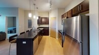 Located at 2900 DeMers Ave in Grand Forks, North Dakota, this single-floor unit offers a well-designed, contemporary living space with a thoughtful layout and modern finishes. The open-concept kitchen and living area serves as the heart of the unit, anchored by a sleek dark granite island with bar seating illuminated by stylish pendant lighting. The kitchen is outfitted with rich dark wood cabinetry, stainless steel appliances including a refrigerator, range, and microwave, complemented by light hardwood-style flooring and track lighting. The adjoining living area provides generous space for seating, with large windows fitted with horizontal blinds that allow ample natural light and outdoor views. Multiple bedrooms feature plush carpeting, natural light via sizable windows, and built-in storage solutions including beds with integrated drawers. Each bathroom is well-appointed with tile flooring, dark vanity cabinetry with granite countertops, a full soaking tub, and built-in wire shelving for added storage. The overall aesthetic is clean and modern, with neutral warm tones, consistent finishes, and functional design elements throughout the entire space.