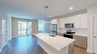 This well-appointed single-floor residence offers a thoughtfully designed open-concept layout combining the kitchen and living room into a bright, cohesive living space. The kitchen is a standout feature, showcasing a large center island with a white quartz countertop and an integrated sink, complemented by white upper cabinetry, warm wood-toned lower cabinets, and a full suite of stainless steel appliances including a refrigerator, electric range, and microwave. Wood-style flooring runs throughout the main living areas, adding warmth and visual continuity. The living room is generously sized and filled with natural light, with sliding glass doors that open directly onto a private balcony overlooking the surrounding building exterior. The bedroom is carpeted for comfort and features a large window that brings in additional natural light. The bathroom is clean and modern, equipped with a single-sink vanity with quartz countertop, wood-toned cabinetry, a glass-enclosed walk-in shower, and wood-style flooring. A dedicated laundry area accommodates a stacked Whirlpool washer and dryer unit alongside built-in shelving for additional storage. The overall design emphasizes a neutral, contemporary aesthetic throughout.