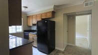 This single-story space showcases a kitchen designed with practicality in mind. The kitchen is equipped with warm-toned wooden cabinetry that provides ample storage, featuring both upper and lower cabinets with raised-panel door detailing. Black appliances, including a full-size refrigerator and a built-in dishwasher, lend a consistent, cohesive look to the space. The dark countertops complement the cabinetry tones, while a single-basin sink is positioned beneath a window, allowing for natural light to enter the workspace. Crown molding runs along the ceiling perimeter, adding a refined architectural detail to the room. The tile flooring in the kitchen offers durability and ease of maintenance. A partial view into an adjacent hallway or room reveals carpeted flooring, suggesting a transition into a living or bedroom area. The neutral wall tones throughout create a clean, understated backdrop that ties the space together. A flush-mount ceiling light provides overhead illumination, keeping the kitchen well-lit. Overall, the space presents a straightforward, functional layout suited for everyday living.