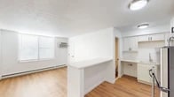 This single-floor unit presents a clean, efficiently designed layout that maximizes its compact footprint. The main living area features warm hardwood-style flooring that extends throughout, crisp white walls, and a textured ceiling. A window unit air conditioner and baseboard heating provide year-round climate control, while a window with horizontal blinds allows natural light to brighten the space.

A white peninsula wall visually separates the living area from the kitchen zone, offering a practical countertop surface. The galley-style kitchen is well-appointed with white shaker cabinets featuring black hardware, light quartz-style countertops, and a full suite of stainless steel appliances including an electric range, over-the-range microwave, and refrigerator. A dedicated sink area with additional cabinetry completes the kitchen.

The bathroom is outfitted with a large walk-in shower enclosed by sliding glass doors with dark framing, a white vanity with under-cabinet storage, and matching hardwood-style flooring that ties into the rest of the unit. The overall aesthetic throughout is consistent and modern, with a neutral white palette that keeps the space feeling bright and move-in ready.
