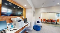 Located at 700 Berkeley Place Circle in Clemson, South Carolina, this well-appointed amenity space serves as a dynamic hub for productivity and collaboration. The single-floor layout is thoughtfully organized into distinct functional zones that flow seamlessly into one another. A dedicated business center area features a built-in wall-mounted desk with a computer workstation and multifunction printer, positioned beneath a large flat-screen TV and an eye-catching geometric gold accent wall. Adjacent to this is a comfortable lounge area furnished with a gray sectional sofa, a bold yellow accent chair, and a vibrant blue tufted ottoman, providing a relaxed setting for informal gatherings. A glass-enclosed conference room offers a more private setting for meetings, outfitted with a long conference table and orange rolling chairs, with natural light filtering through its windows. The main lounge area also features a round white meeting table surrounded by coordinating orange chairs, set against large windows that invite ample natural light and outdoor views. Throughout the space, contemporary artwork, patterned carpet tile flooring, recessed lighting, and carefully curated furnishings in orange, yellow, and blue create an energetic and welcoming atmosphere ideal for residents and professionals alike.