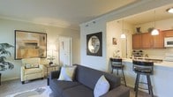 This single-story residence showcases an open-concept design that seamlessly connects the living area to the kitchen. The living room is comfortably proportioned, featuring crown molding along the ceiling that adds a refined architectural detail. Neutral wall tones create a warm, welcoming ambiance, complemented by soft carpeting underfoot. A charcoal gray sofa serves as the focal point of the seating area, accompanied by a classic armchair and a side table with a table lamp that provides warm accent lighting. A framed circular mirror in a bold black square frame adds a decorative element to the main wall.

The kitchen is visible through an open pass-through, which enhances the sense of spaciousness and allows for easy interaction between the two areas. A pendant light hangs above a breakfast bar equipped with bar-height seating, making it a practical spot for casual dining. The kitchen features warm wood cabinetry and what appears to be light-colored countertops. A white paneled door leads to an additional room, contributing to the functional and well-connected layout of this residence.