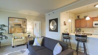 This single-story residence showcases an open-concept design that seamlessly connects the living area to the kitchen. The living room is comfortably proportioned, featuring crown molding along the ceiling that adds a refined architectural detail. Neutral wall tones create a warm, welcoming ambiance, complemented by soft carpeting underfoot. A charcoal gray sofa serves as the focal point of the seating area, accompanied by a classic armchair and a side table with a table lamp that provides warm accent lighting. A framed circular mirror in a bold black square frame adds a decorative element to the main wall.

The kitchen is visible through an open pass-through, which enhances the sense of spaciousness and allows for easy interaction between the two areas. A pendant light hangs above a breakfast bar equipped with bar-height seating, making it a practical spot for casual dining. The kitchen features warm wood cabinetry and what appears to be light-colored countertops. A white paneled door leads to an additional room, contributing to the functional and well-connected layout of this residence.