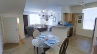 This single-floor residence offers a thoughtfully designed open-concept layout that seamlessly connects the main living areas. The dining area serves as a central focal point, anchored by an elegant chandelier with candle-style lighting that hangs above a round glass-top dining table — a classic yet refined touch. Light-toned walls and flooring carry throughout, creating a bright, cohesive aesthetic.

Adjacent to the dining space, the kitchen features warm wood cabinetry, dark countertops, and a peninsula-style breakfast bar that provides additional seating and a natural transition between the kitchen and living areas. The kitchen is well-appointed with standard appliances and ample cabinet storage.

The living room, visible from the dining area, benefits from generous natural light streaming through large windows. Comfortable seating, including a sofa and accent chairs, gives the space a welcoming, relaxed character. A ceiling fan overhead adds both function and casual style.

The open flow between all three areas — dining, kitchen, and living room — makes this home well-suited for everyday living and entertaining. The consistent use of neutral tones and classic furnishings throughout lends the space a timeless, livable quality.