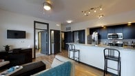 This single-floor residence offers a thoughtfully designed open-concept layout that seamlessly connects the living, dining, and kitchen areas. The living space is anchored by warm hardwood flooring and features a ceiling fan, creating a comfortable and inviting atmosphere. Bold dark-painted doors and trim provide a striking contrast against the light walls, lending a contemporary edge to the interior design.

The kitchen is a standout feature, showcasing rich dark cabinetry paired with elegant granite countertops that extend into a breakfast bar — ideal for casual dining or entertaining. A full suite of stainless steel appliances, including a refrigerator, electric range, and built-in microwave, ensures both style and functionality. Track lighting above the kitchen provides focused illumination, complementing the recessed lighting throughout.

Multiple doorways visible from the main living area suggest well-proportioned additional rooms, including what appears to be a bedroom and bathroom, each featuring the same consistent dark door styling. The overall ambiance is modern and cohesive, with a neutral color palette accented by dark tones, creating a space that feels both sophisticated and practical.