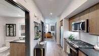 This efficiently designed one-bedroom, one-bathroom unit maximizes its compact footprint with a thoughtful, linear layout and contemporary finishes throughout. Light hardwood-style flooring runs continuously across the entire space, creating visual cohesion and an airy feel.

The galley-style kitchen is a standout feature, outfitted with warm wood-tone cabinetry, dark stone countertops, a white penny-tile backsplash, and a full suite of stainless steel appliances including a gas range, built-in microwave, and refrigerator. An entry corridor lined with open shelving provides a stylish transition into the living and sleeping areas.

The generously sized bedroom accommodates a full seating and workspace arrangement alongside the bed, demonstrating strong versatility. A glass door opens directly to an outdoor area with lush greenery, bringing in natural light and a refreshing connection to the outdoors. Built-in shelving adds functional storage and visual interest.

The bathroom continues the unit's clean aesthetic with light flooring, crisp white walls, and refined fixtures. Throughout, the unit's modern, carefully curated design makes efficient use of every square foot.