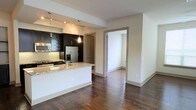 This single-story Dallas space showcases a thoughtfully designed open-concept layout that seamlessly connects the kitchen and living areas. The kitchen is a standout feature, outfitted with rich, dark cabinetry that provides a striking contrast against light granite countertops. A peninsula with an integrated sink extends into the living space, offering both additional prep space and a natural division between the two areas. Stainless steel appliances, including a French door refrigerator and gas range, lend a modern, professional feel to the kitchen. Pendant lighting hangs above the peninsula, adding warm, focused illumination that complements the recessed track lighting overhead. Dark hardwood floors run throughout, grounding the space with warmth and a cohesive visual flow. Neutral-toned walls keep the interior feeling bright and open. A hallway visible from the main living area leads to additional rooms, suggesting a functional and well-organized floor plan. The overall design balances contemporary aesthetics with practical living, making it a polished and inviting space well-suited for both everyday living and entertaining.